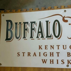 May include: A white banner with the word "BUFFALO" in large, dark green letters with a brown outline. Below, the text reads "KENTUCKY STRAIGHT BOURBON WHISKEY". A brown bison graphic is on the right.