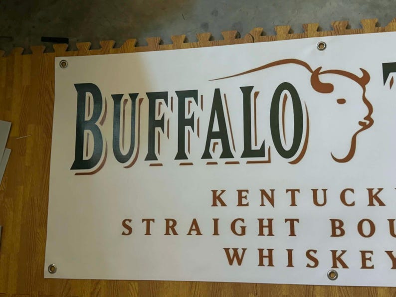 May include: A white banner with the word "BUFFALO" in large, dark green letters with a brown outline. Below, the text reads "KENTUCKY STRAIGHT BOURBON WHISKEY". A brown bison graphic is on the right.