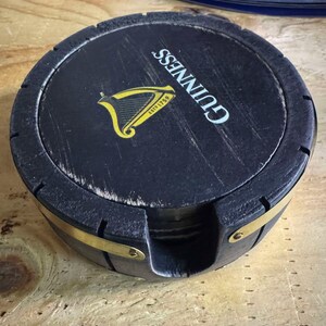 May include: A set of four black wooden coasters with a brass band around the edge. The top coaster features the Guinness logo with the text "GUINNESS" and "ESTD 1759" in yellow.