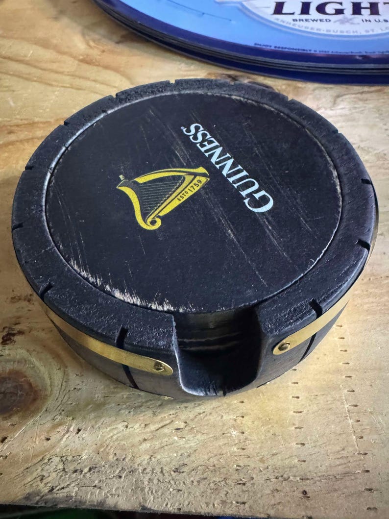 May include: A set of four black wooden coasters with a brass band around the edge. The top coaster features the Guinness logo with the text "GUINNESS" and "ESTD 1759" in yellow.