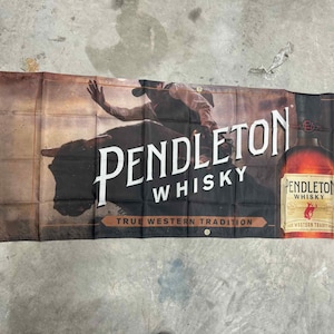 Peut inclure: Bannière Pendleton Whisky avec une silhouette de cow-boy et un graphisme de bouteille. La bannière présente le nom de la marque en grandes lettres blanches, avec le slogan "True Western Tradition". La palette de couleurs est marron, noir et blanc.