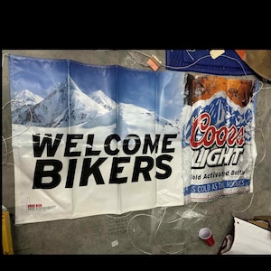 Könnte beinhalten: Ein weißes Banner mit dem Text "WELCOME BIKERS" in großen schwarzen Buchstaben. Das Banner zeigt eine Berglandschaft und ein Coors Light Bierdosen-Design. Das Coors Light Dosen-Design enthält den Text "Coors Light" und "Cold Activated Bottle".