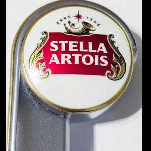 Pode incluir: Close-up de uma torneira de cerveja Stella Artois. O emblema circular apresenta o logotipo Stella Artois em vermelho e branco, com detalhes dourados e uma estrela. A torneira é prateada.
