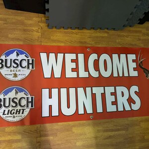 May include: Red banner with the words "WELCOME HUNTERS" in white block letters. The banner also features the Busch Beer and Busch Light logos, along with a deer graphic.