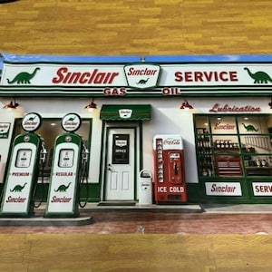 May include: A rectangular wall sign depicting a vintage Sinclair gas station. The scene features two green gas pumps, a red Coca-Cola ice machine, a white office door, and a service bay window. The Sinclair logo with a green dinosaur is visible.