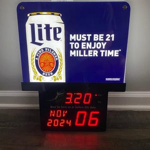 May include: A blue and black sign featuring a Miller Lite beer can graphic and the text "MUST BE 21 TO ENJOY MILLER TIME." Below is a digital clock displaying the time, date, and the text "Must be born on or before this date."
