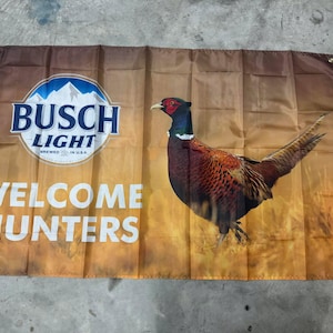 Può includere: Uno stendardo marrone e dorato con il logo Busch Light e il testo "Welcome Hunters". Al centro dello stendardo è raffigurato un fagiano.