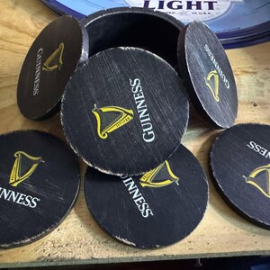May include: Six black round coasters with the word "GUINNESS" printed in white and a gold harp logo. The coasters are arranged in a circle around a wooden box.
