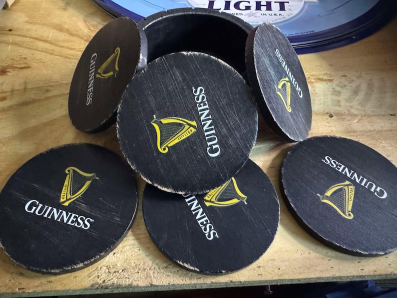 May include: Six black round coasters with the word "GUINNESS" printed in white and a gold harp logo. The coasters are arranged in a circle around a wooden box.
