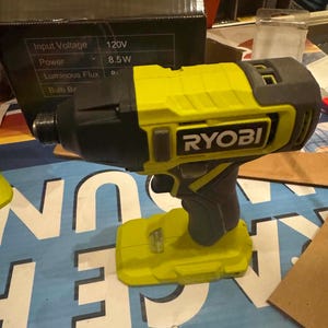 Könnte beinhalten: Ein neongrüner und schwarzer Ryobi Akku-Schlagschrauber mit Batteriebasis. Das Werkzeug verfügt über einen schwarzen Griff, einen Abzug und den auf der Seite aufgedruckten Markennamen Ryobi. Es steht vor einem schwarzen Karton mit technischen Daten.
