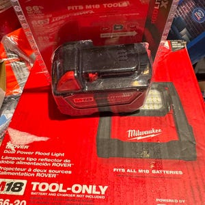 May include: A black Milwaukee M18 REDLITHIUM XC5.0 battery in its original plastic packaging, resting on a red cardboard box for a Milwaukee ROVER Dual Power Flood Light that states it fits all M18 batteries and is tool-only.