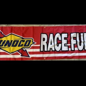 May include: A red banner with white stripes, displaying the text "SUNOCO RACE FUELS." The Sunoco logo is in the centre, a yellow diamond with a blue outline and the word "SUNOCO" in blue.