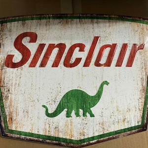 Puede incluir: Letrero de gasolinera Sinclair vintage con un acabado envejecido. El letrero presenta el nombre Sinclair en letras rojas en bloque sobre una silueta de dinosaurio verde sobre un fondo blanco. El letrero tiene un borde verde.