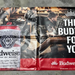 May include: A banner featuring a Budweiser beer can and a mechanic working on a motorcycle. The banner has the text "THIS BUD'S FOR YOU" in large white letters. The Budweiser logo is also visible.