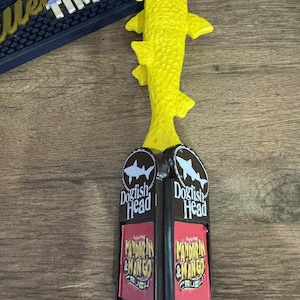 Könnte beinhalten: Ein gelber, fischförmiger Zapfhahn mit dem Text "Dogfish Head" und "Mandarin & Mango Crush" auf einem schwarz-roten Hintergrund. Der Zapfhahn besteht aus einem strukturierten Material.