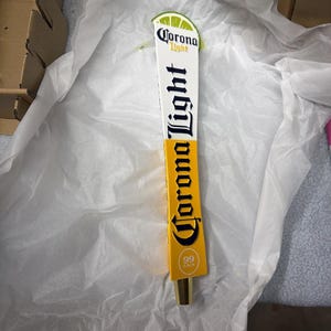 May include: A vertical Corona Light beer tap handle featuring a white top section with the brand logo and a yellow bottom section with the text Corona Light and 99 Cals. The top is accented with a green lime wedge graphic.