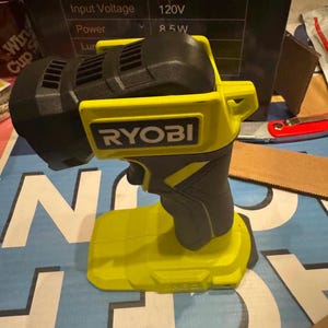 Könnte beinhalten: Eine limettengrüne und schwarze Ryobi LED-Handarbeitsleuchte mit Abzugsschalter, schwenkbarem Kopf und flacher Basis für Stabilität. Das Gerät zeigt den Markennamen Ryobi an der Seite und den eingeprägten Text ONE+ auf der Basis des Werkzeugs.