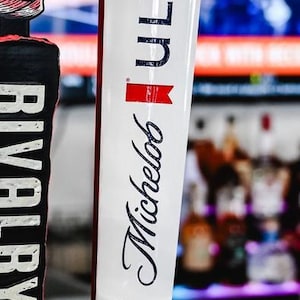 May include: A white beer tap handle with the word "ULTRA" in dark blue capital letters. Below, the word "Michelob" is written in a cursive font. A red and white logo is also visible. The tap handle is on a bar.