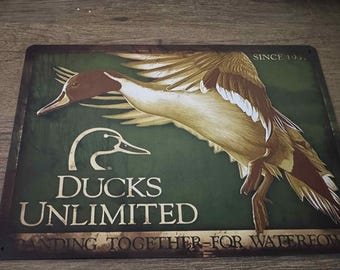 Może przedstawiać: Prostokątny metalowy znak z zielonym tłem przedstawiający szczegółową ilustrację lecącej kaczki. Znak zawiera tekst "DUCKS UNLIMITED" i "STANDING TOGETHER FOR WATERFOWL" z napisem "SINCE 1937" na górze.