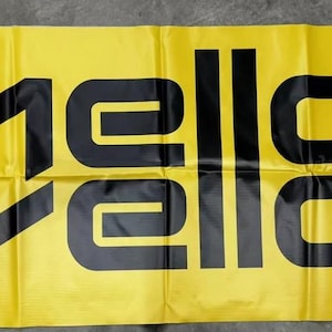 Puede incluir: Una pancarta amarilla con el texto "Mello Yello" en letras negras. El "Yello" está en una fuente estilizada con una flecha negra apuntando hacia abajo. La pancarta tiene un símbolo de marca registrada en la esquina inferior derecha.