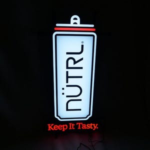May include: A white and red illuminated sign featuring the word "NUTRL" in black, stylized text. The sign is shaped like a beverage container, with the text "Keep It Tasty" in red below it, against a black backdrop.