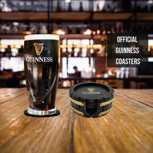 May include: Set of four black and gold Guinness coasters. The coasters are shaped like a barrel and have the Guinness logo on them. The coasters are sitting on a wooden table next to a glass of Guinness.