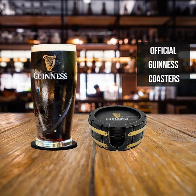 May include: Set of four black and gold Guinness coasters. The coasters are shaped like a barrel and have the Guinness logo on them. The coasters are sitting on a wooden table next to a glass of Guinness.