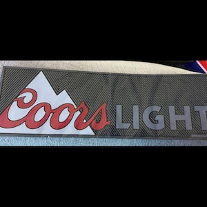 以下が含まれることがあります： Coors Lightのロゴが入った長方形のバーマット。ロゴには、赤い筆記体で「Coors」の文字と白い山のグラフィック、そしてグレーで「LIGHT」の文字が含まれています。マットは、テクスチャ加工された濃いグレーの背景です。