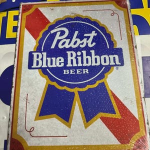 May include: A vintage Pabst Blue Ribbon Beer metal sign. The sign features a blue ribbon design with the brand name in white lettering. The background is white with red and gold accents, giving it a retro look.