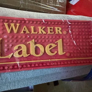 May include: A red bar mat with the text "JOHNNIE WALKER Label" in gold lettering. The mat has a textured surface with small, raised dots. The product is wrapped in clear plastic.