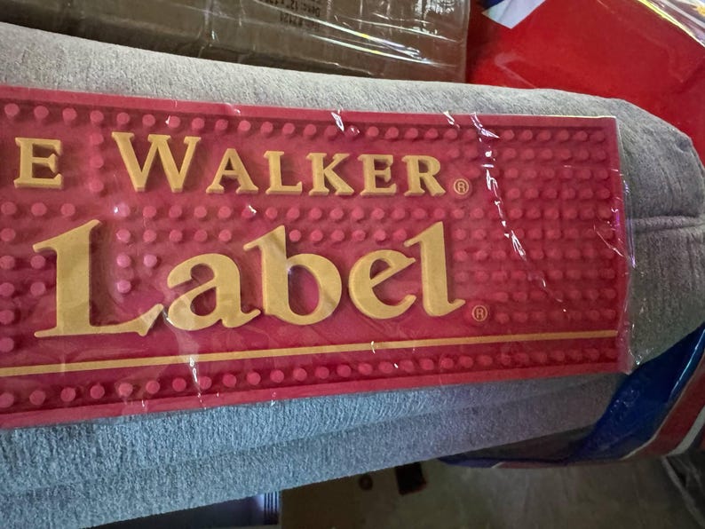 May include: A red bar mat with the text "JOHNNIE WALKER Label" in gold lettering. The mat has a textured surface with small, raised dots. The product is wrapped in clear plastic.