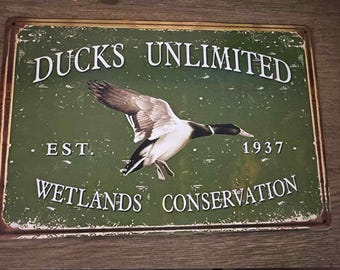 Może przedstawiać: Prostokątny metalowy znak z postarzanym wykończeniem. Znak zawiera tekst "DUCKS UNLIMITED" nad wizerunkiem lecącej kaczki. Poniżej tekst "WETLANDS CONSERVATION" i rok "1937". Znak jest zielony i brązowy.