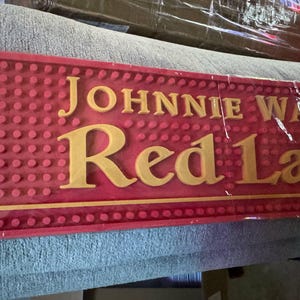 May include: A red bar mat with the words "JOHNNIE WALKER Red Label" in gold lettering. The mat has a textured surface with small, raised dots. The mat is rectangular and appears to be made of rubber.