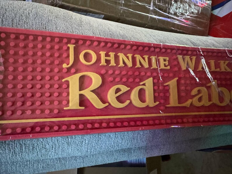 May include: A red bar mat with the words "JOHNNIE WALKER Red Label" in gold lettering. The mat has a textured surface with small, raised dots. The mat is rectangular and appears to be made of rubber.