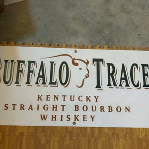 May include: A white banner with the words "BUFFALO TRACE" in large, dark green letters with a brown outline. Below, it reads "KENTUCKY STRAIGHT BOURBON WHISKEY" in brown. A bison graphic is featured.