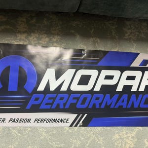 May include: A rectangular vinyl banner featuring the blue Mopar logo and the white text MOPAR PERFORMANCE. The bottom section includes the text POWER. PASSION. PERFORMANCE. in black on a white background, with metal grommets in the corners.
