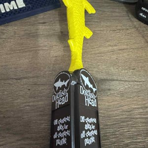 Peut inclure: Un robinet de bière en forme de poisson jaune avec une base rectangulaire noire et blanche. La base présente le logo "Dogfish Head" et le texte "Off-centered ales for off-centered people."