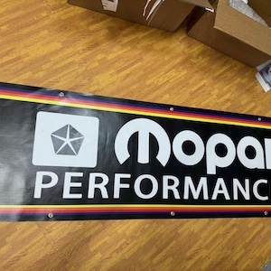 May include: Black rectangular sign with the white text "Mopar PERFORMANCE" and a white pentagon logo. The sign has a yellow, red, and blue border. The sign is mounted with silver rivets.