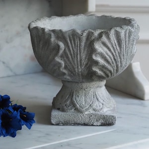 Könnte beinhalten: Grauer, verwitterter Beton-Pflanztopf mit Sockel und gewelltem Rand. Der Pflanztopf hat ein detailliertes Blattdesign und steht auf einer Marmoroberfläche. Ein kleiner Strauß blauer Blumen befindet sich im Vordergrund.