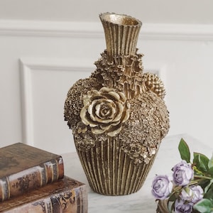 Könnte beinhalten: Eine goldfarbene dekorative Vase mit strukturierter Oberfläche und Rosendesign. Die Vase hat einen geriffelten Hals und ist mit floralen und strukturierten Elementen verziert. Die Vase steht auf einer Marmoroberfläche.