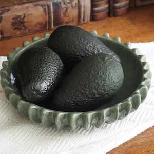 Könnte beinhalten: Drei reife Avocados liegen in einer salbeigrünen Keramikschale mit gewelltem Rand. Die Schale steht auf einer weißen, strukturierten Oberfläche mit einem hölzernen Hintergrund. Die Avocados haben eine dunkelgrüne, strukturierte Schale.