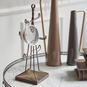 Può includere: Un espositore per tazze in metallo color bronzo con manico attorcigliato, che tiene una tazza da tè a fantasia floreale. La tazza da tè ha uno sfondo bianco con un motivo floreale. Altri elementi decorativi includono vasi e libri.