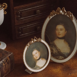 Peut inclure: Deux cadres ovales anciens avec des bordures dorées et un accent de perle. Les portraits représentent des femmes en costumes d'époque. Une coupe décorative et un livre vintage sont également visibles sur une surface en bois.