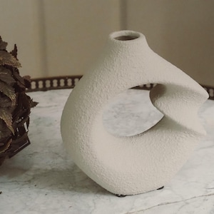 Peut inclure: Un vase en céramique texturée de couleur crème avec un design ouvert unique. Le vase a une base arrondie, une ouverture centrale et une forme sculpturale torsadée. La surface a une finition mate rugueuse. Décoration intérieure.