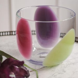 Op de afbeelding: Een helder glazen tumbler met drie abstracte, kleurrijke vormen: roze, paars en lichtgroen. Het glas staat op een decoratief dienblad met een donkere rand, met een paarse bloem en groene bladeren op de voorgrond.
