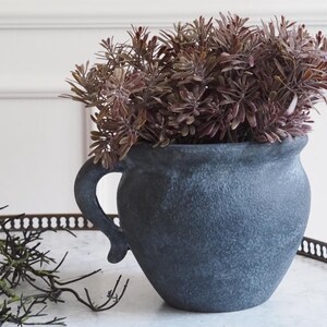 Può includere: Un vaso in ceramica blu scuro e strutturato con manico, riempito con fogliame finto marrone-rossastro. Il vaso ha un aspetto rustico e invecchiato e poggia su un vassoio decorativo. Adatto per l'arredamento della casa.