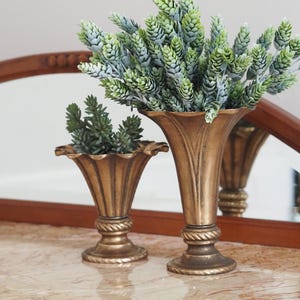 Könnte beinhalten: Zwei goldene Metallvasen mit geriffeltem Design. Die größere Vase ist mit einem Strauß grüner Kunstblumen gefüllt. Die kleinere Vase ist leer.