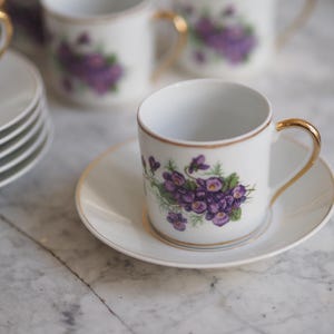Könnte beinhalten: Weißes Porzellan-Teeservice mit einem Blumendesign aus lila Veilchen und grünen Blättern. Die Tasse hat einen goldenen Henkel und Zierrat. Mehrere Tassen und Untertassen sind sichtbar, was auf ein Teeservice hindeutet.