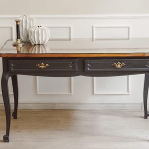Peut inclure: Un bureau gris anthracite foncé avec des pieds galbés et deux tiroirs dotés de poignées en métal doré. Le plateau présente un bord en bois naturel et un dessus en verre transparent. Deux vases en céramique blanche texturée et un bougeoir sont posés.