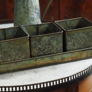 Könnte beinhalten: Drei quadratische Metallpflanzgefäße mit geprägten Blumenmustern stehen auf einem passenden rechteckigen Tablett. Das Set hat eine verwitterte grün-bronze Oberfläche, die einen Vintage-Stil suggeriert. Die Pflanzgefäße und das Tablett stehen auf einem Tisch mit Marmorplatte.